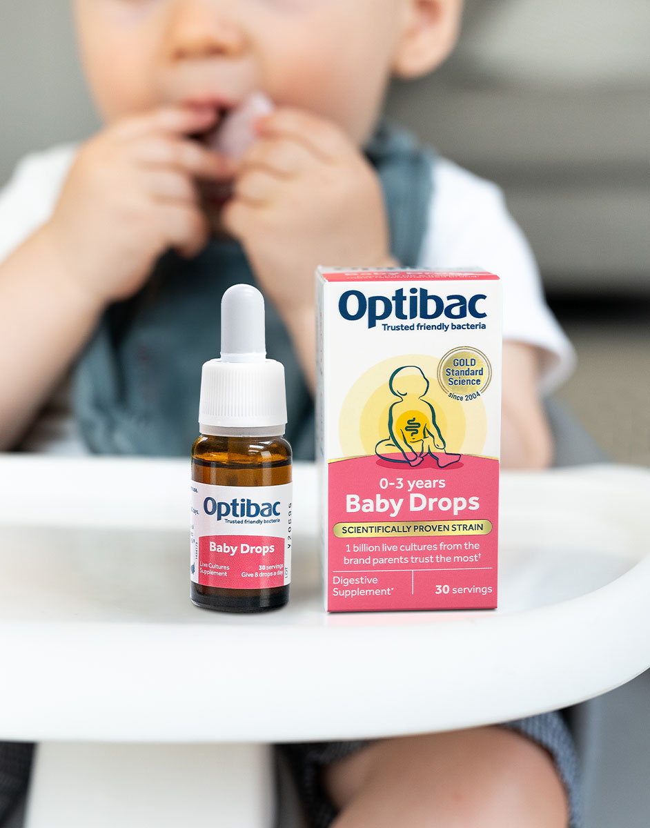 Baby holding a dropper bottle of Optibac Baby Drops, with the product box beside it on a high chair tray.
