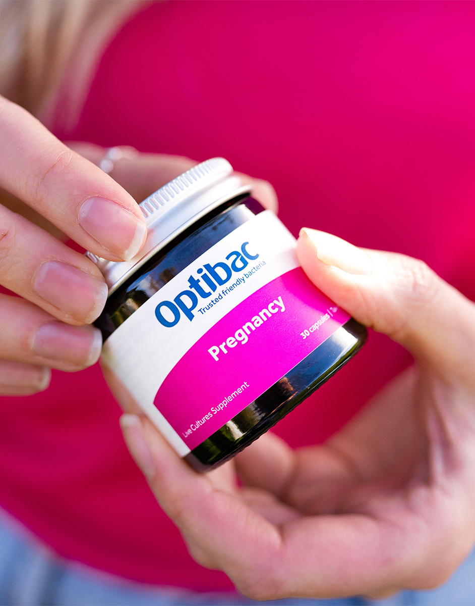 Hands holding a jar of Optibac Pregnancy probiotic supplement against a pink background.