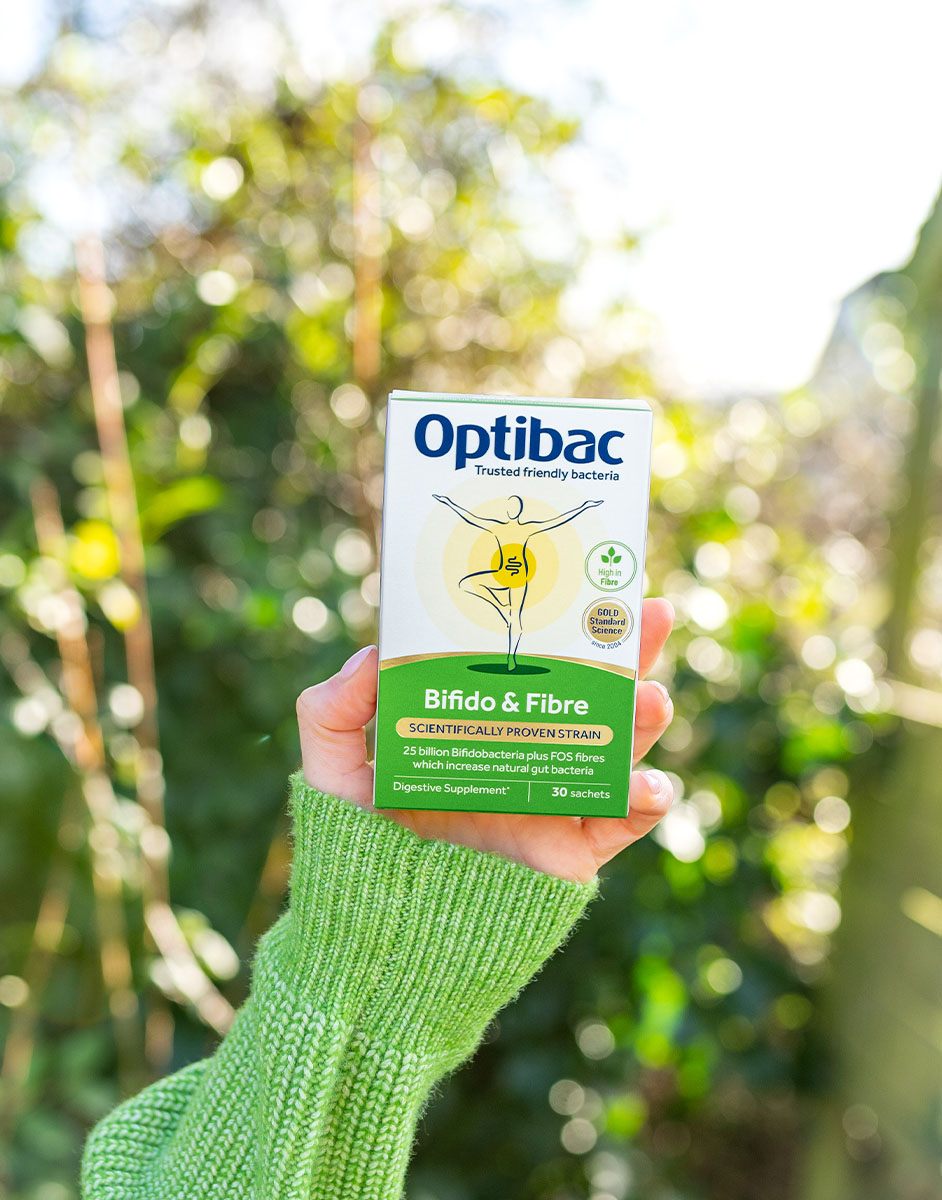 A hand in a green sweater holds a box of Optibac Bifido & Fibre sachets against a blurred outdoor background.