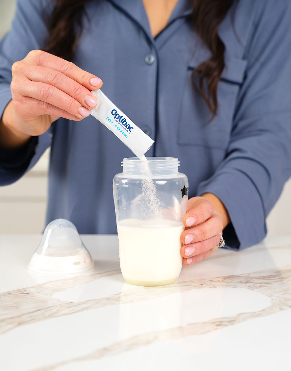 Person adding a sachet of Optibac probiotics to a baby bottle filled with milk on a marble countertop.