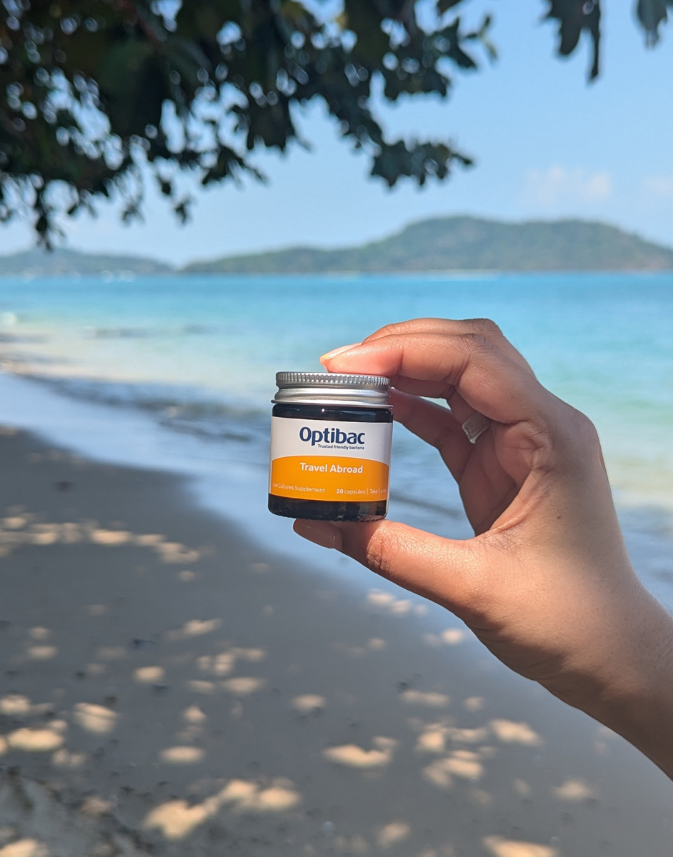 Hand holding a jar of Optibac Travel Abroad probiotics on a sunny beach with turquoise water and distant islands in the background.