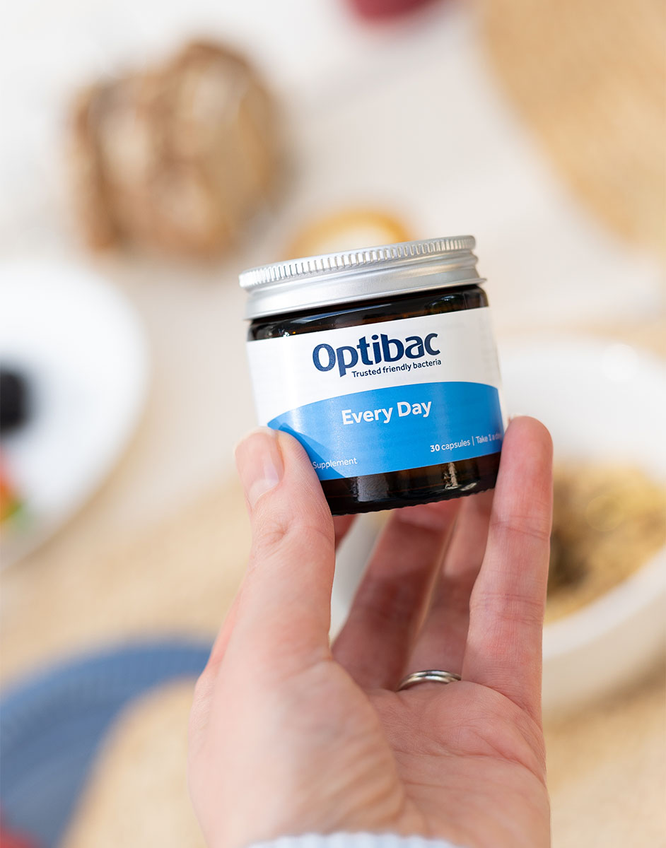 Hand holding a jar of Optibac Every Day probiotics with a blurred background of a table setting.