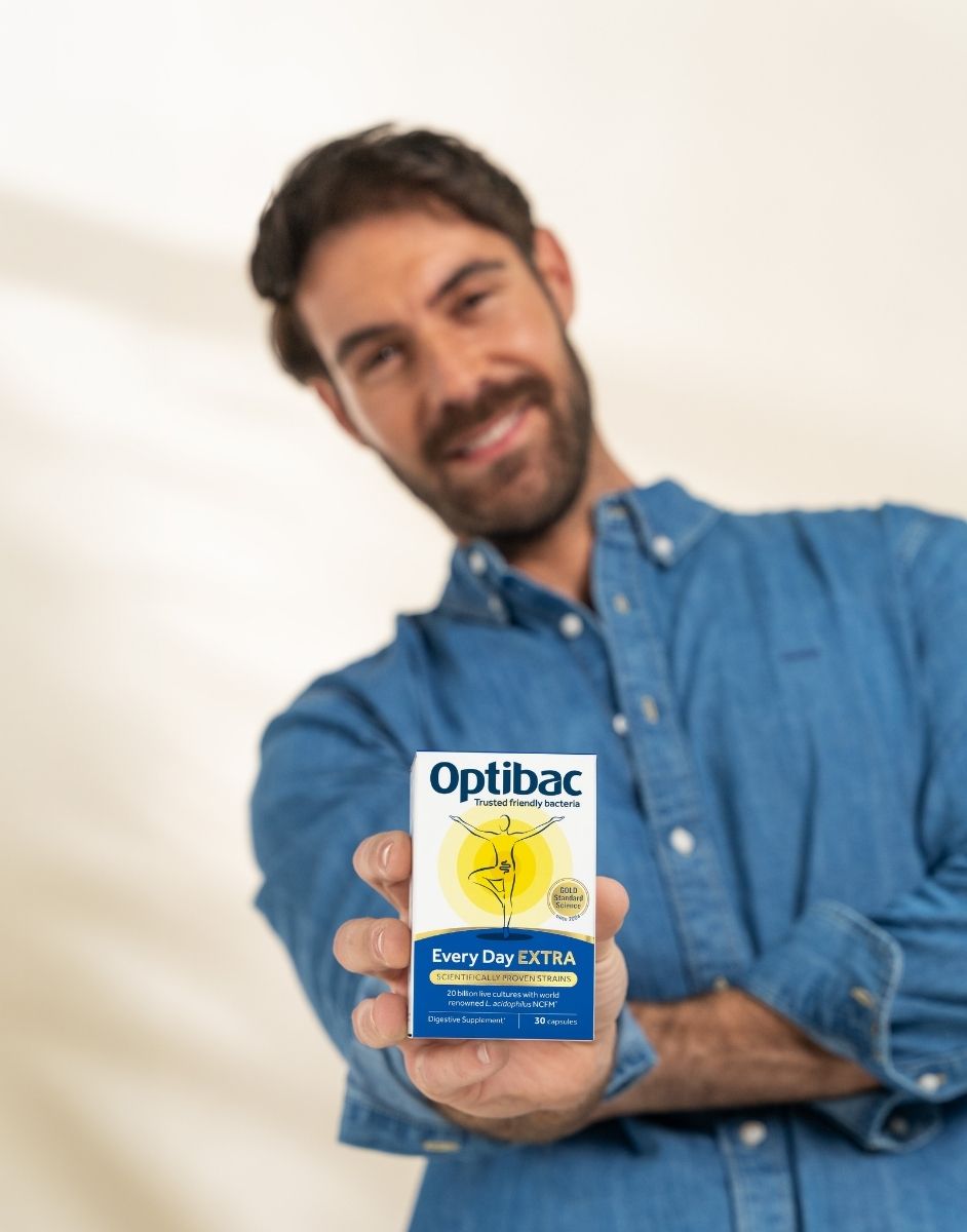 Man in denim shirt holding a box of Optibac Every Day EXTRA probiotics towards the camera, smiling against a neutral background.