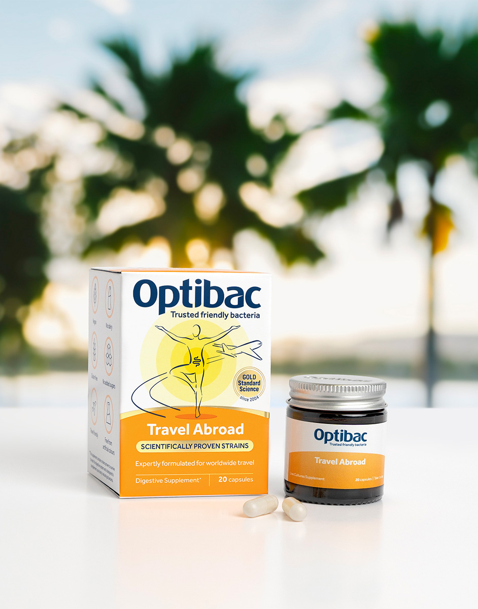 Optibac Travel Abroad supplement box and jar with capsules on a table, palm trees blurred in the background.