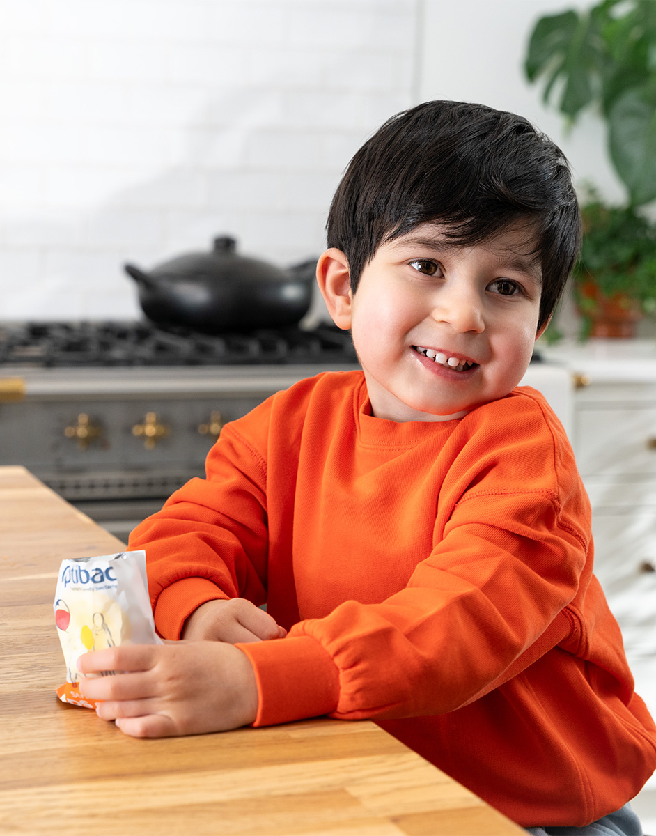 Smiling child in an orange sweater holds a snack pack at a kitchen table, with a stove and plant in the background.