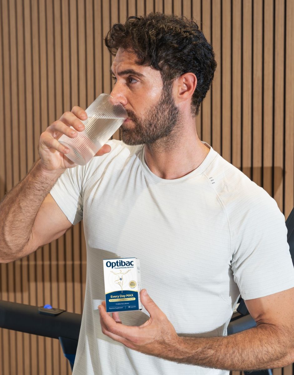 A man in a white shirt drinks water while holding a box of Optibac Every Day MAX probiotics, standing in front of a wooden wall.