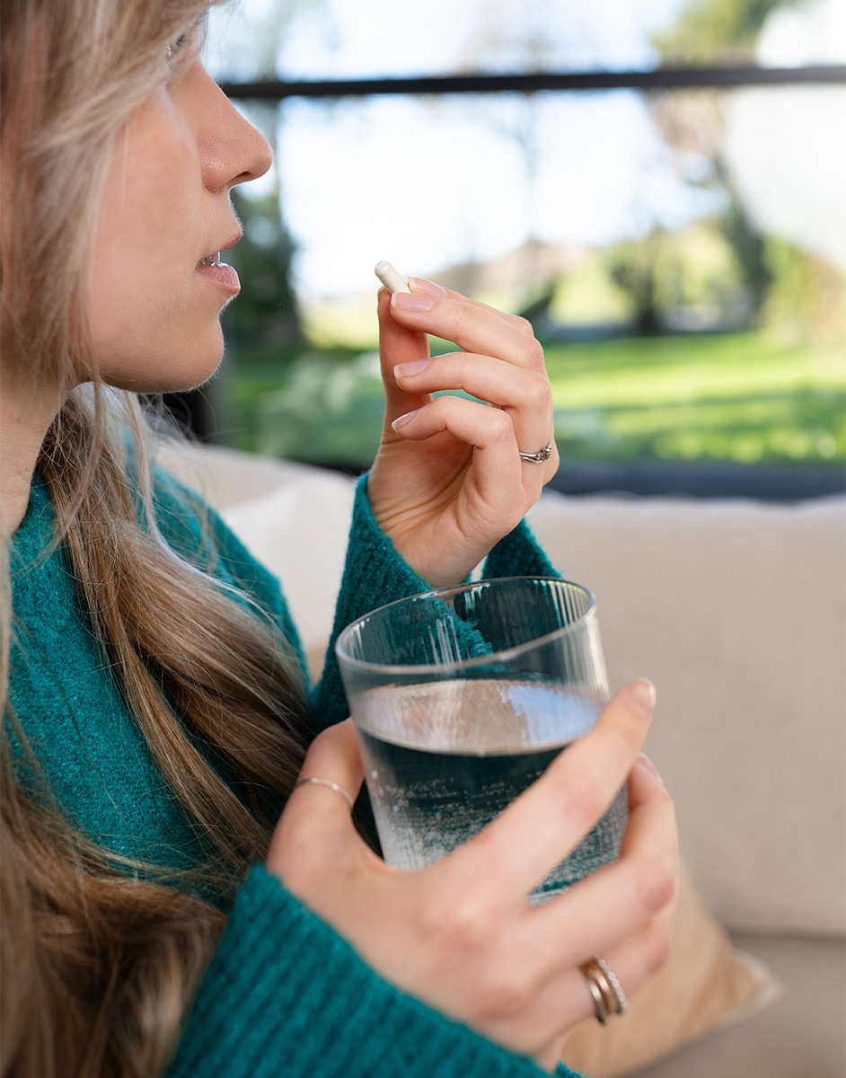 A person in a teal sweater holds a pill and a glass of water, sitting indoors with a view of a garden outside.