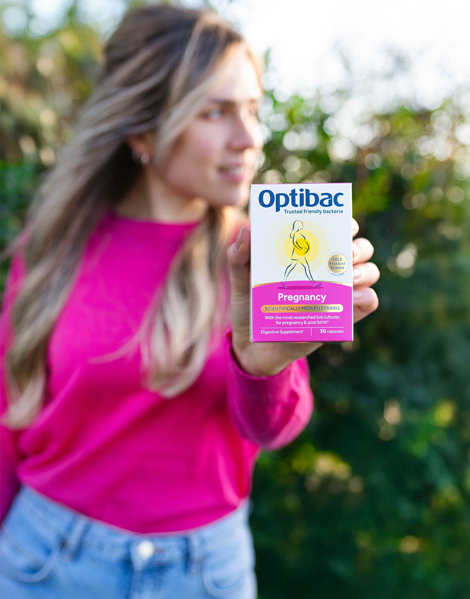 Person in a pink shirt holds a box of Optibac Pregnancy supplements outdoors, with greenery in the background.