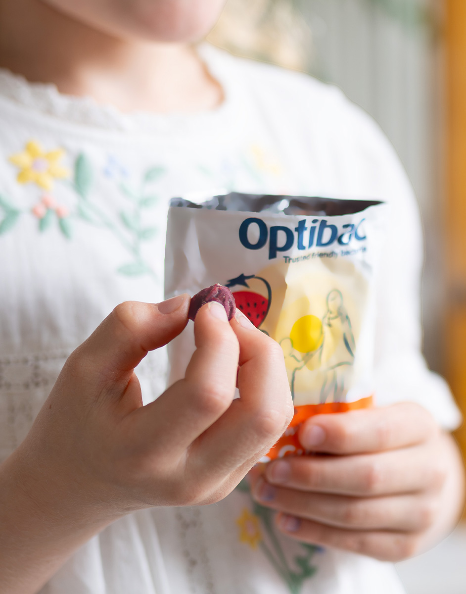Child in a white floral dress holding a packet of Optibac and a small, round item, possibly a supplement or candy.