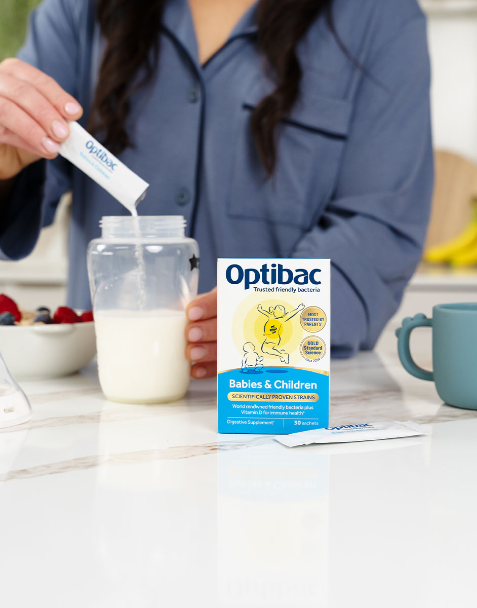 Person mixing Optibac probiotic sachet into a jar of milk on a kitchen counter, with a box labeled "Babies & Children" in the foreground.