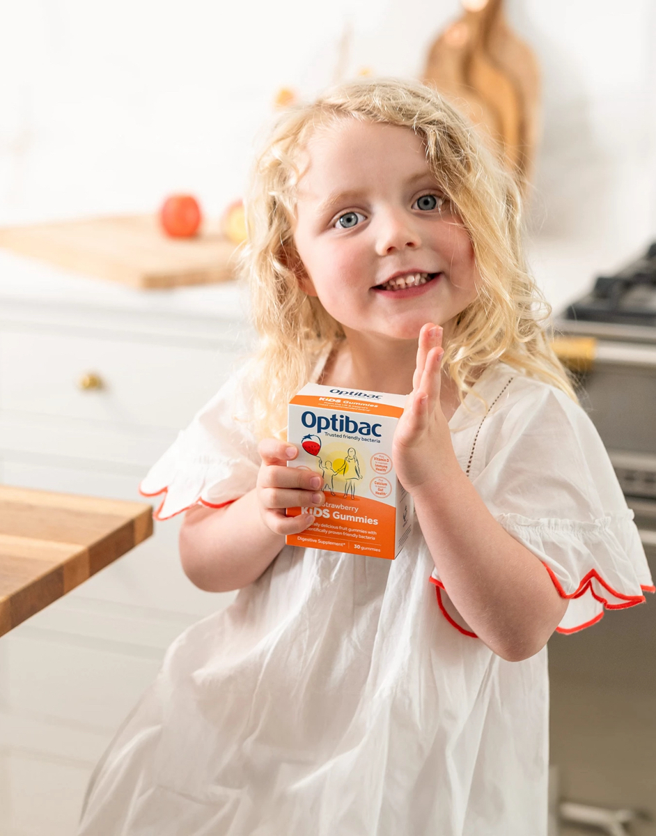 Young girl with curly blonde hair smiling and holding a box of Optibac Kids Gummies in a bright kitchen setting.