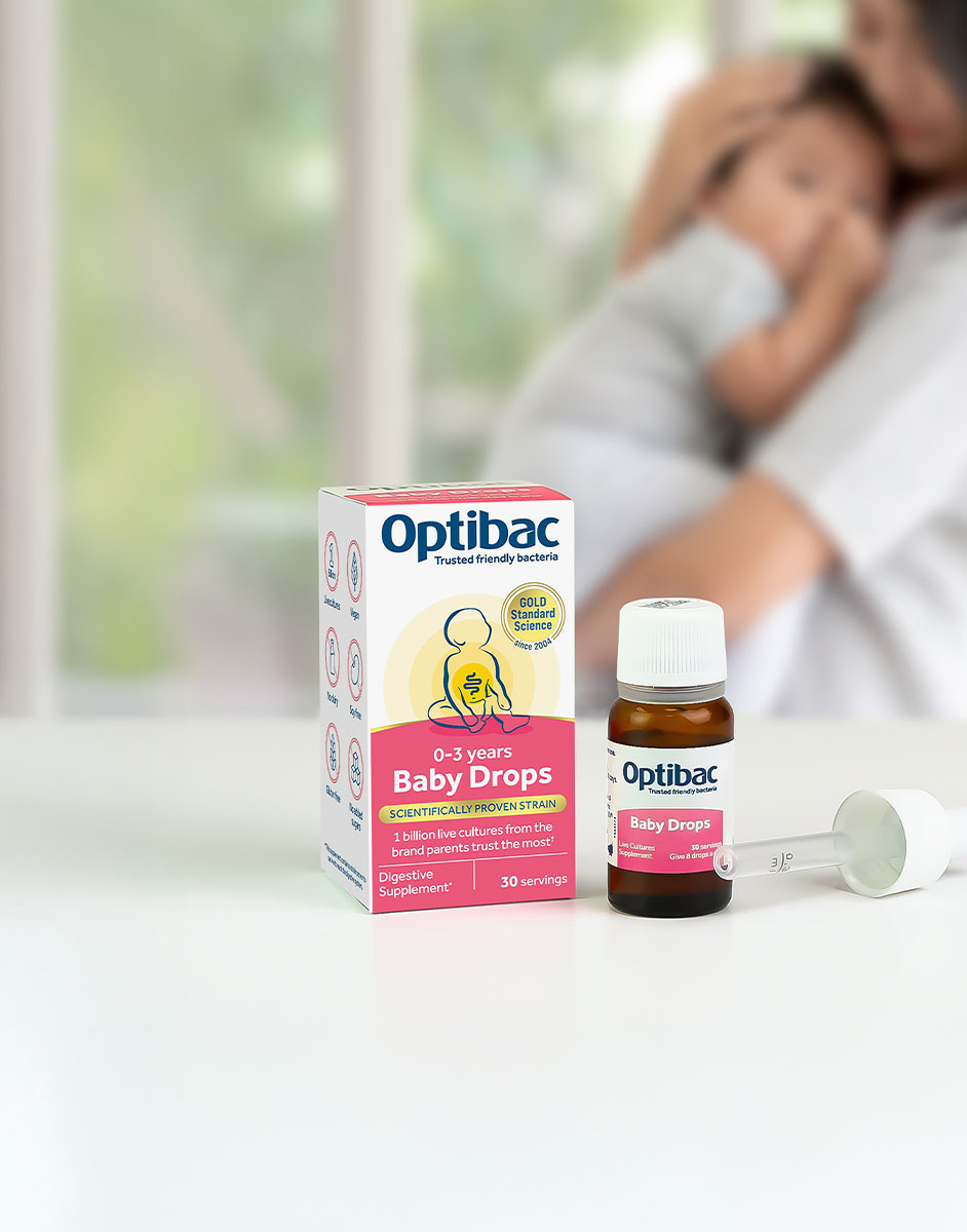 Optibac Baby Drops bottle and box on a table, with a dropper. A person holds a baby in the blurred background.