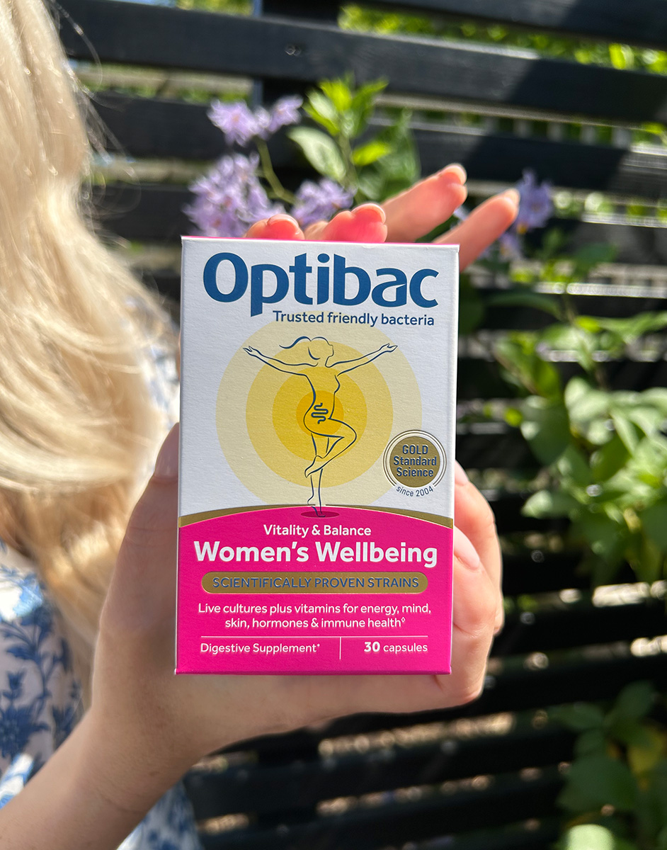 A person holds a box of Optibac Women's Wellbeing supplements outdoors, with purple flowers in the background.