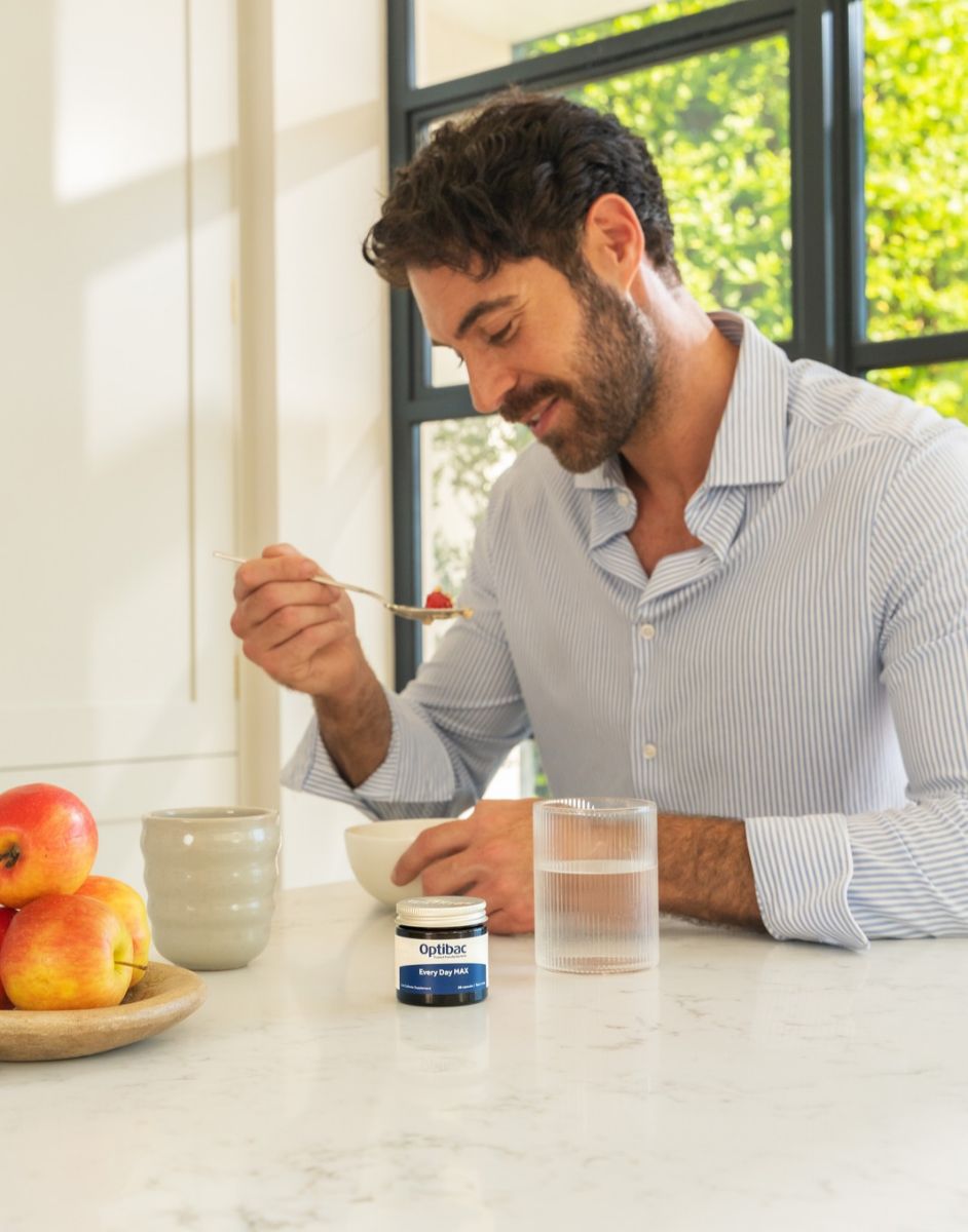 A man in a striped shirt sits at a kitchen table, eating with a spoon. Nearby are apples and a jar labeled "Optibac Every Day MAX."