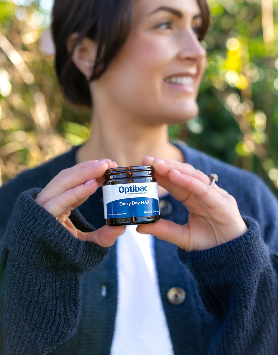 Person holding a jar of Optibac Every Day MAX outdoors, smiling and looking away.