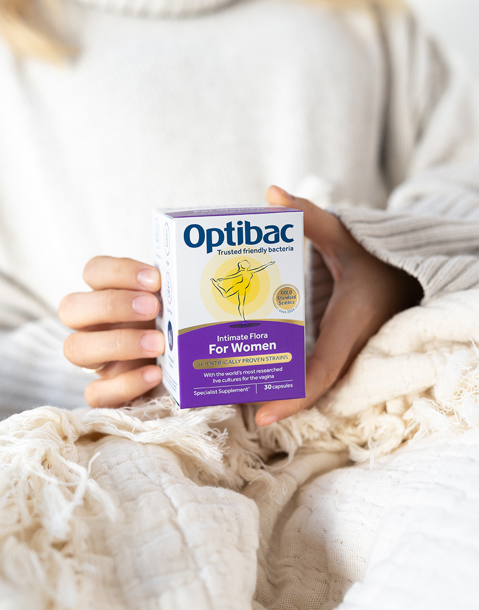 A person in a cozy sweater holds a box of Optibac Intimate Flora for Women, a probiotic supplement, against a soft, textured blanket.
