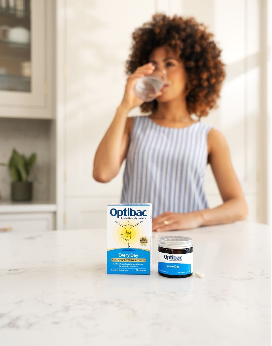 A woman in a striped shirt drinks a glass of water in the background, with Optibac probiotic packaging and a jar in the foreground on a countertop.