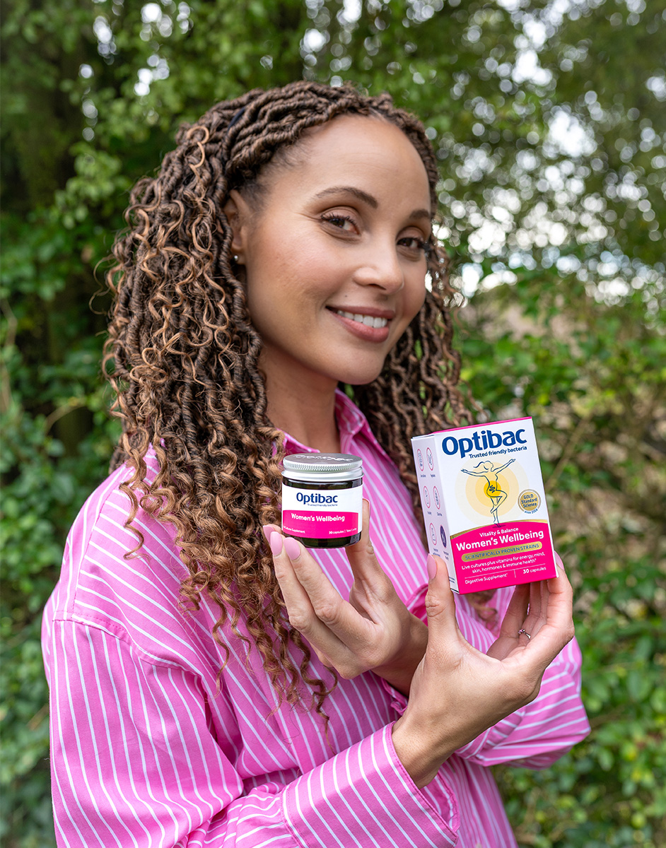 Woman in a pink shirt holding Optibac Women's Wellbeing supplement box and jar, smiling outdoors with greenery in the background.