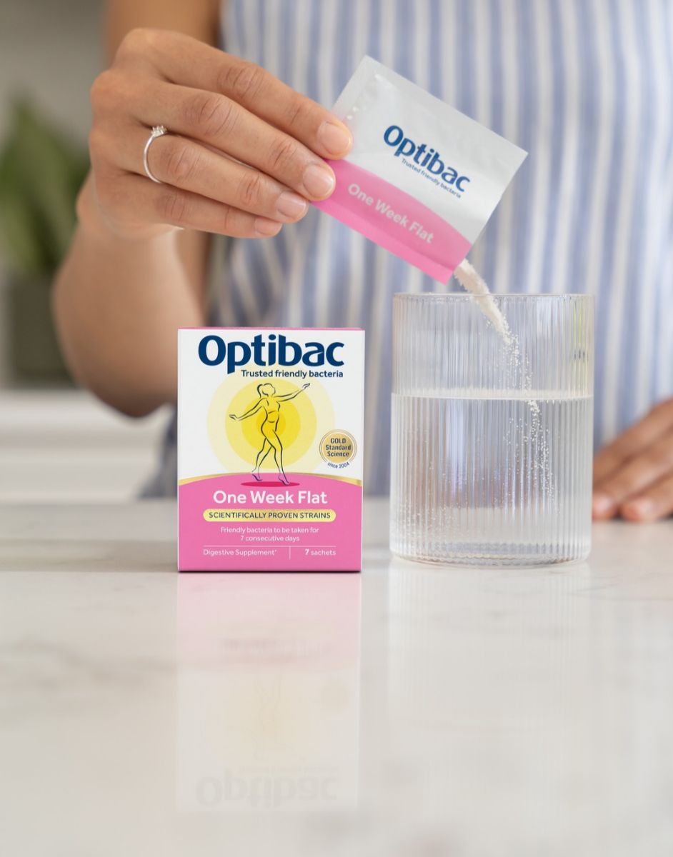 A person pours a sachet of Optibac One Week Flat probiotic powder into a glass of water next to the product box on a countertop.