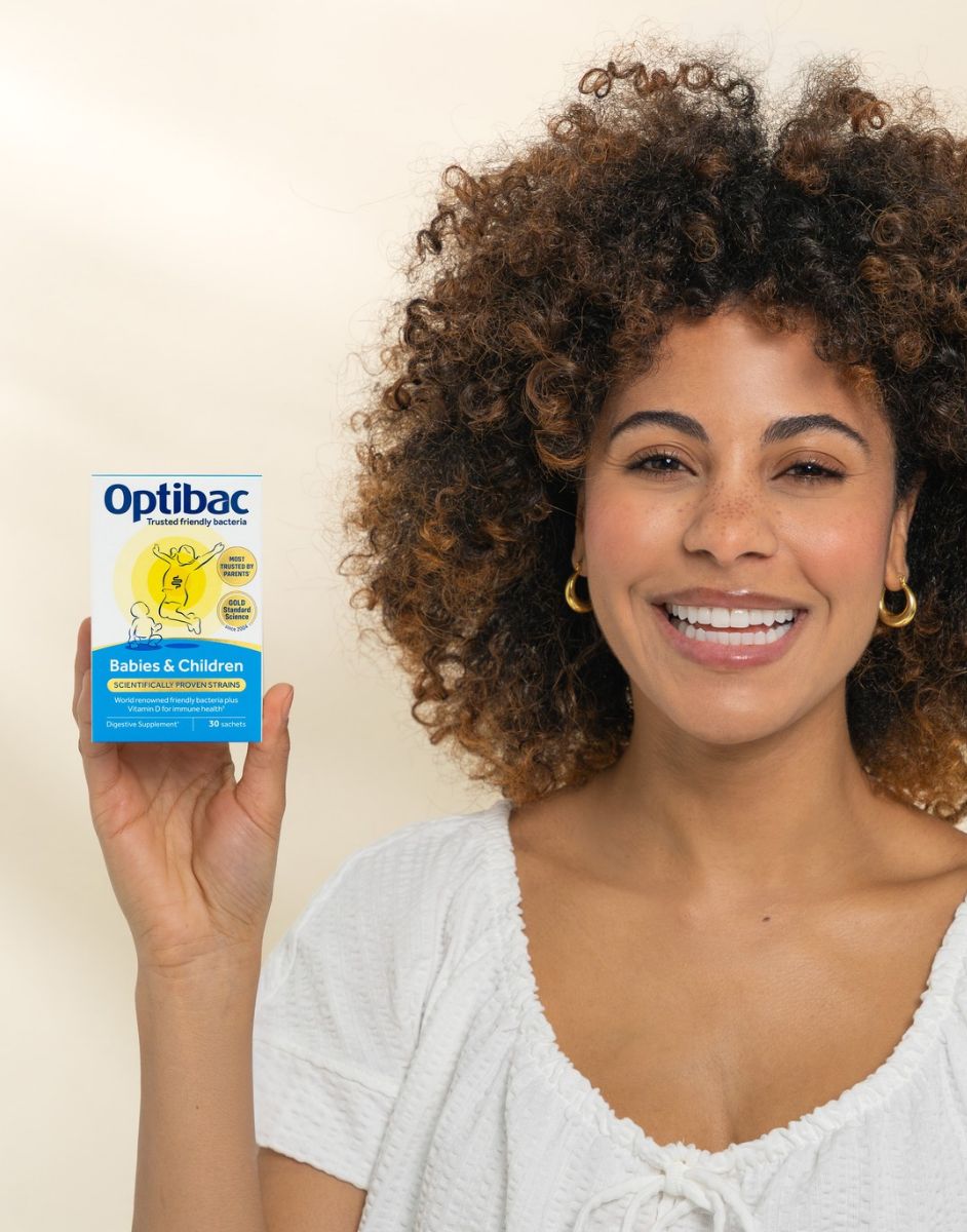 Smiling woman with curly hair holds a box of Optibac probiotics for babies and children, wearing a white top and gold hoop earrings.