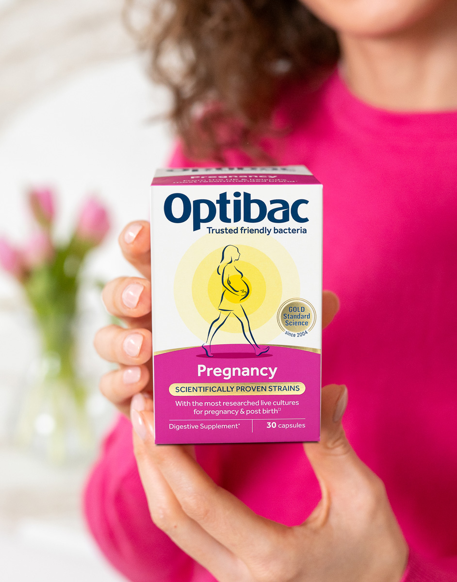 A person in a pink sweater holds a box of Optibac Pregnancy supplements, featuring a pregnant woman illustration and text about probiotic strains.