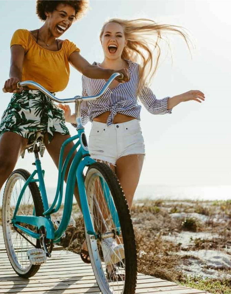 Two women joyfully ride a blue bicycle on a sunny boardwalk, with one peddling and the other balancing beside her, both laughing.