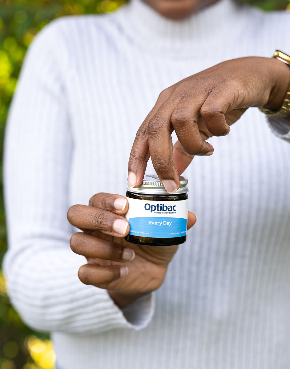 Person in a gray sweater opening a jar of Optibac Every Day supplements outdoors.