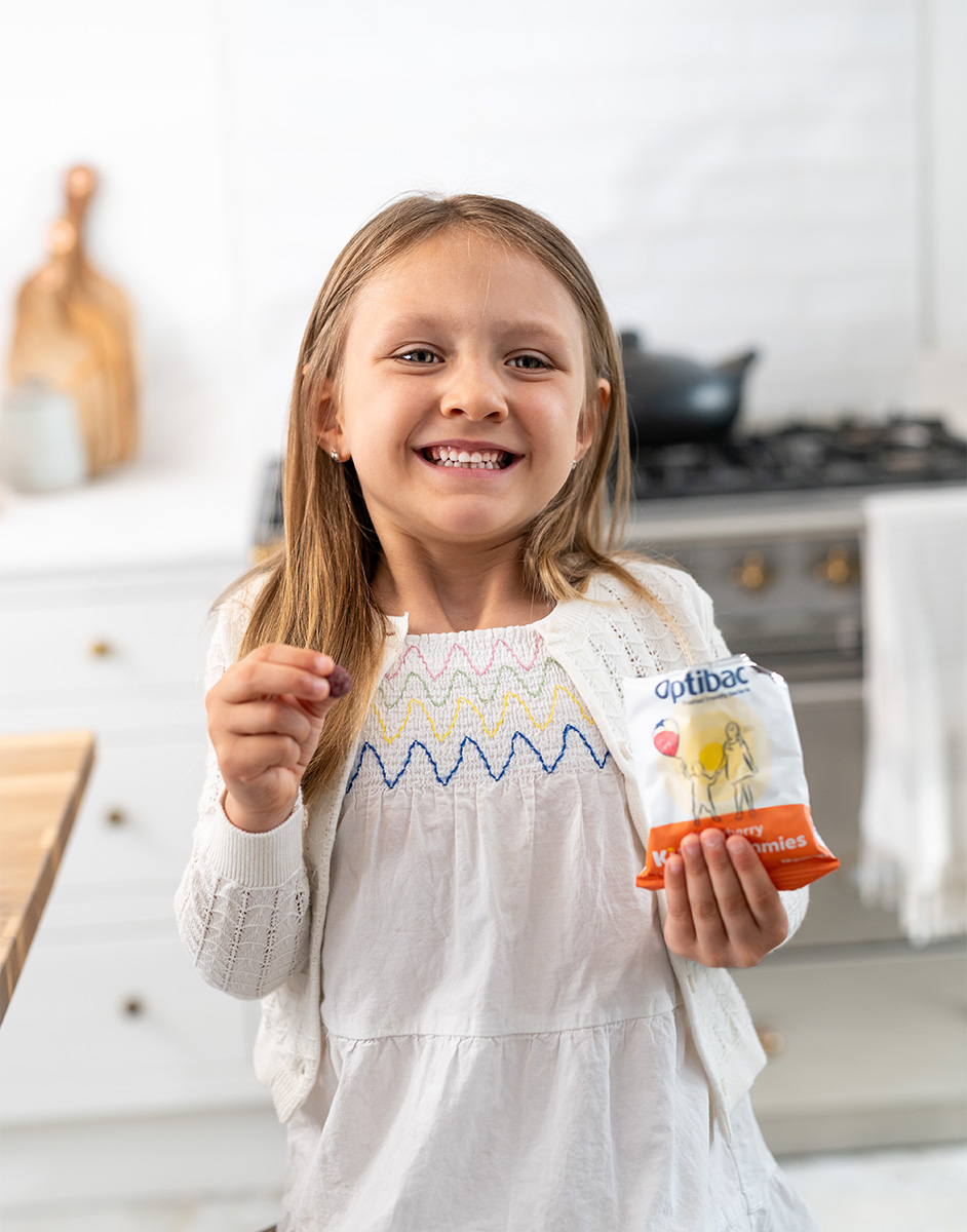 Smiling girl in a kitchen holding a packet labeled "Optibac." She wears a white dress with colorful patterns and a cardigan.
