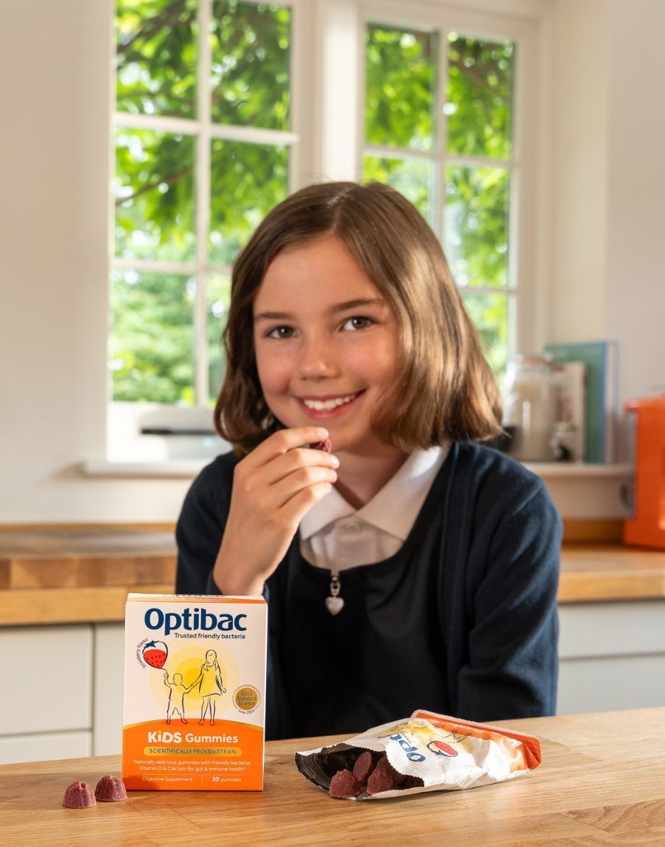 A child in a kitchen holding a gummy, with a box and pack of Optibac Kids Gummies on the table in front of her.