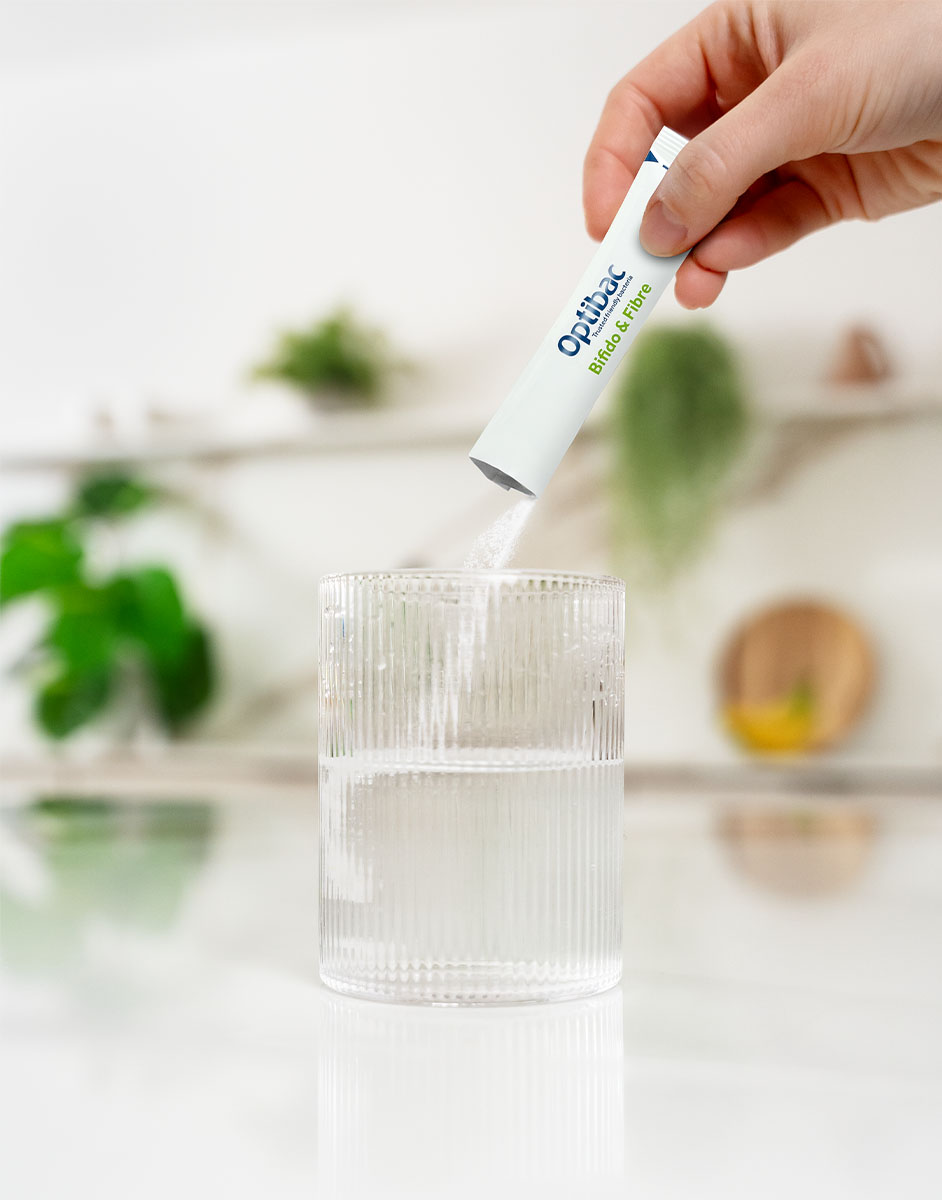 A hand pours a sachet of Optibac Probiotics powder into a clear glass of water on a white countertop with plants in the background.