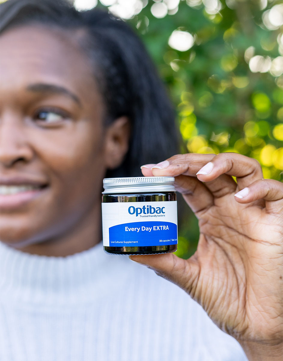 Person holding a jar of Optibac Every Day EXTRA supplements, with a blurred green background.