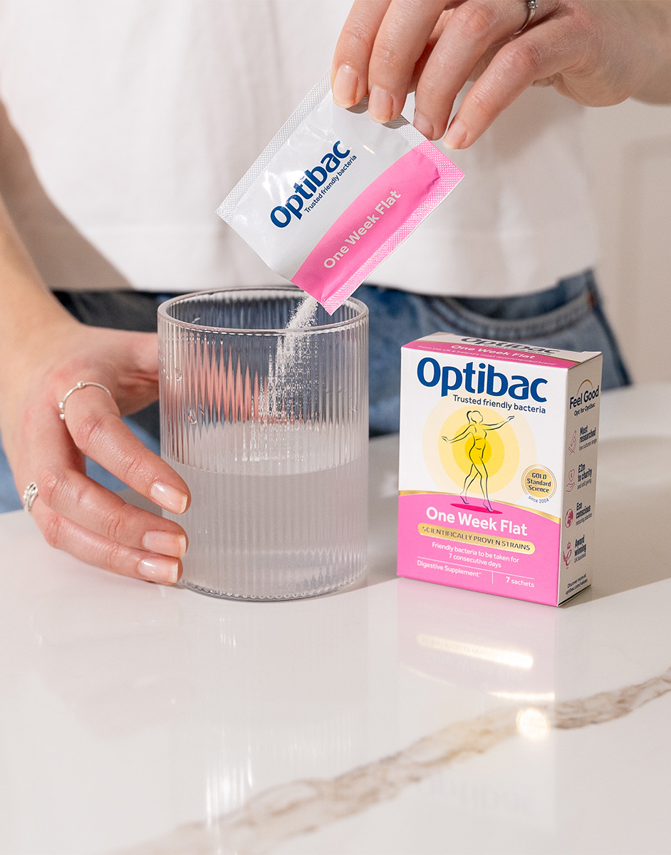 Person pouring Optibac probiotic powder into a glass of water, with product packaging visible on a white countertop.