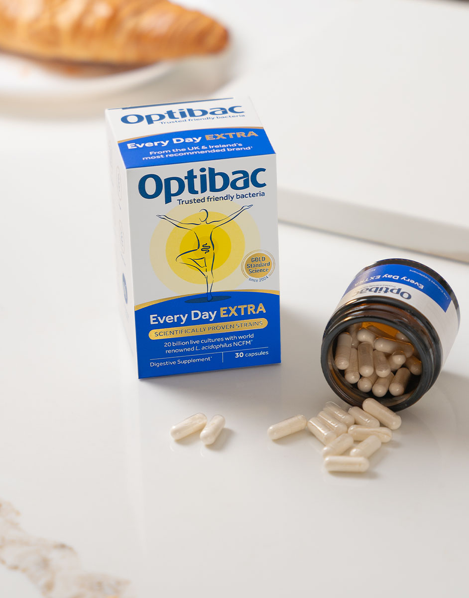 Optibac Every Day EXTRA probiotic box and open bottle with capsules on a white surface, with a blurred croissant in the background.