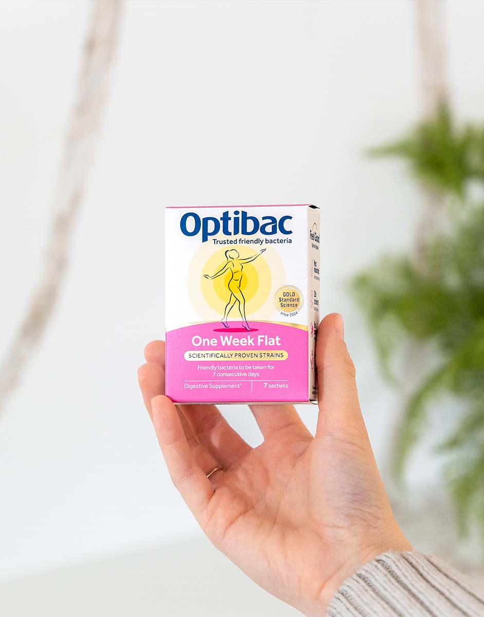 Hand holding a box of Optibac One Week Flat digestive supplement with a pink and white design, against a blurred background.