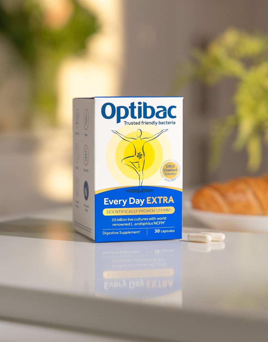 Box of Optibac Every Day EXTRA probiotics with two capsules beside it on a table, blurred background with plants and bread.