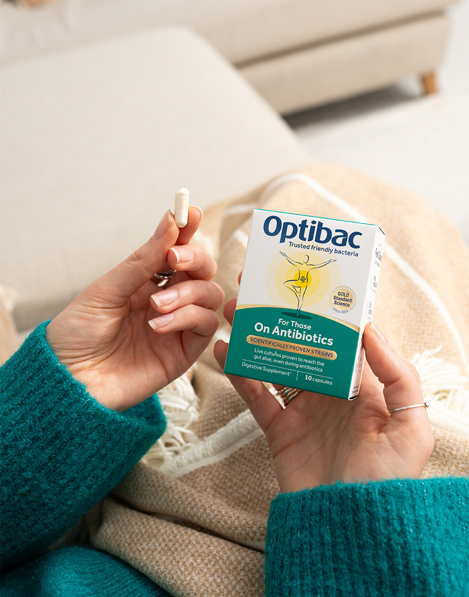 Person holding a probiotic capsule and an Optibac box labeled "For Those On Antibiotics," sitting on a beige and white blanket.