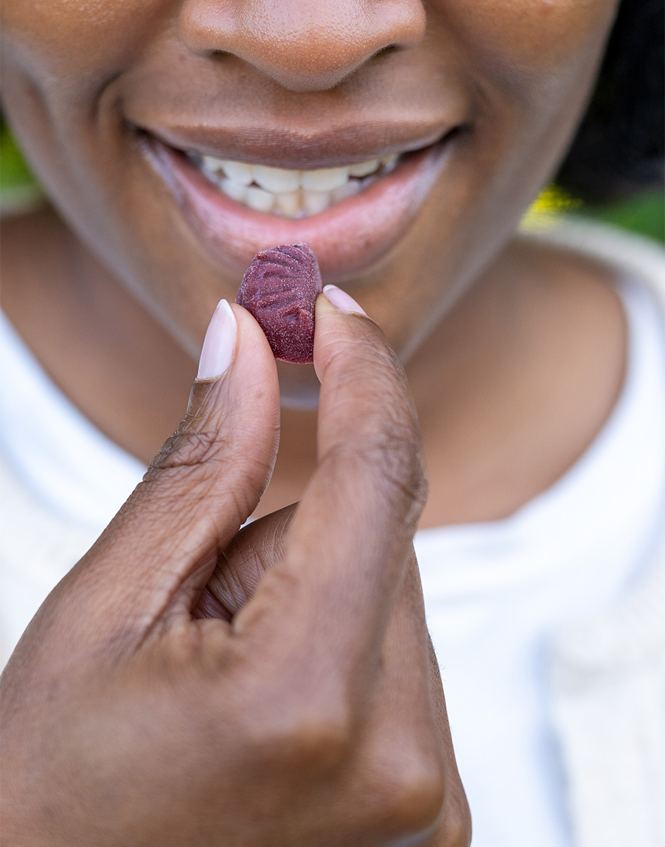 Person smiling while holding a small, red gummy near their lips, ready to eat. Close-up of hands and mouth, with a blurred background.