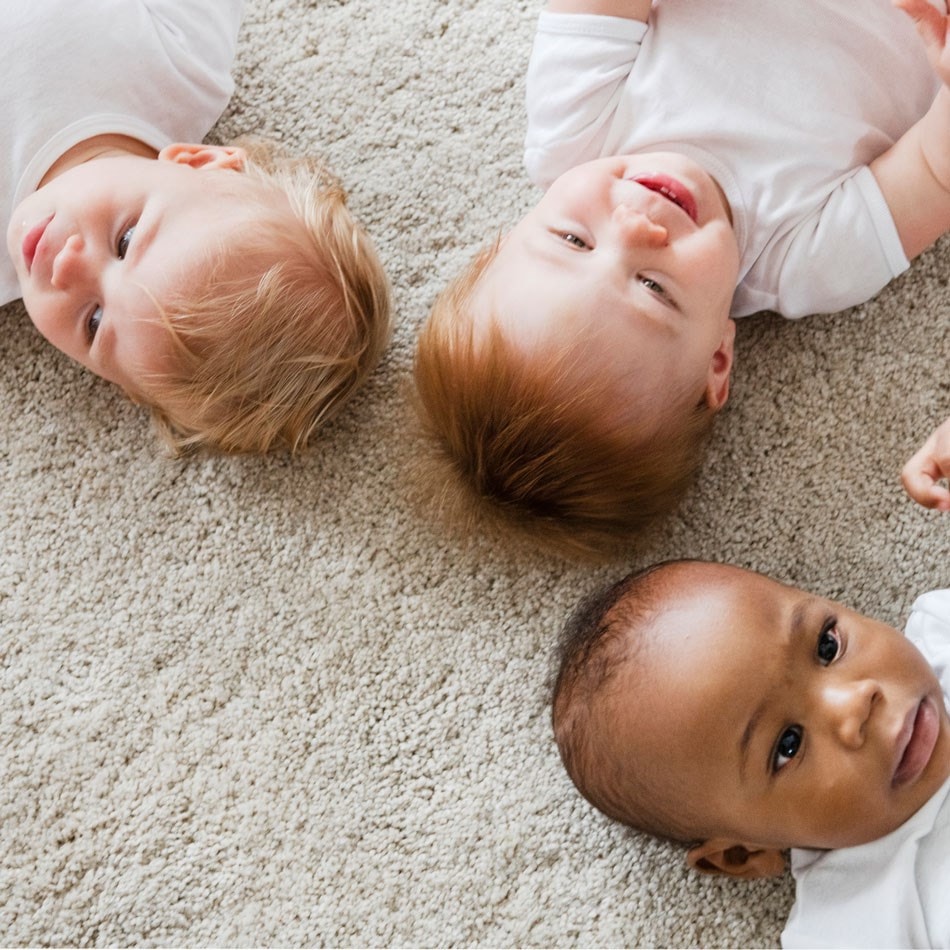 Three babies in white shirts lying on a soft, beige carpet, looking up and smiling.