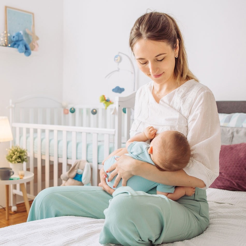 A mother is sitting on a bed, breastfeeding her baby in a cozy, softly lit nursery with a crib and toys in the background.