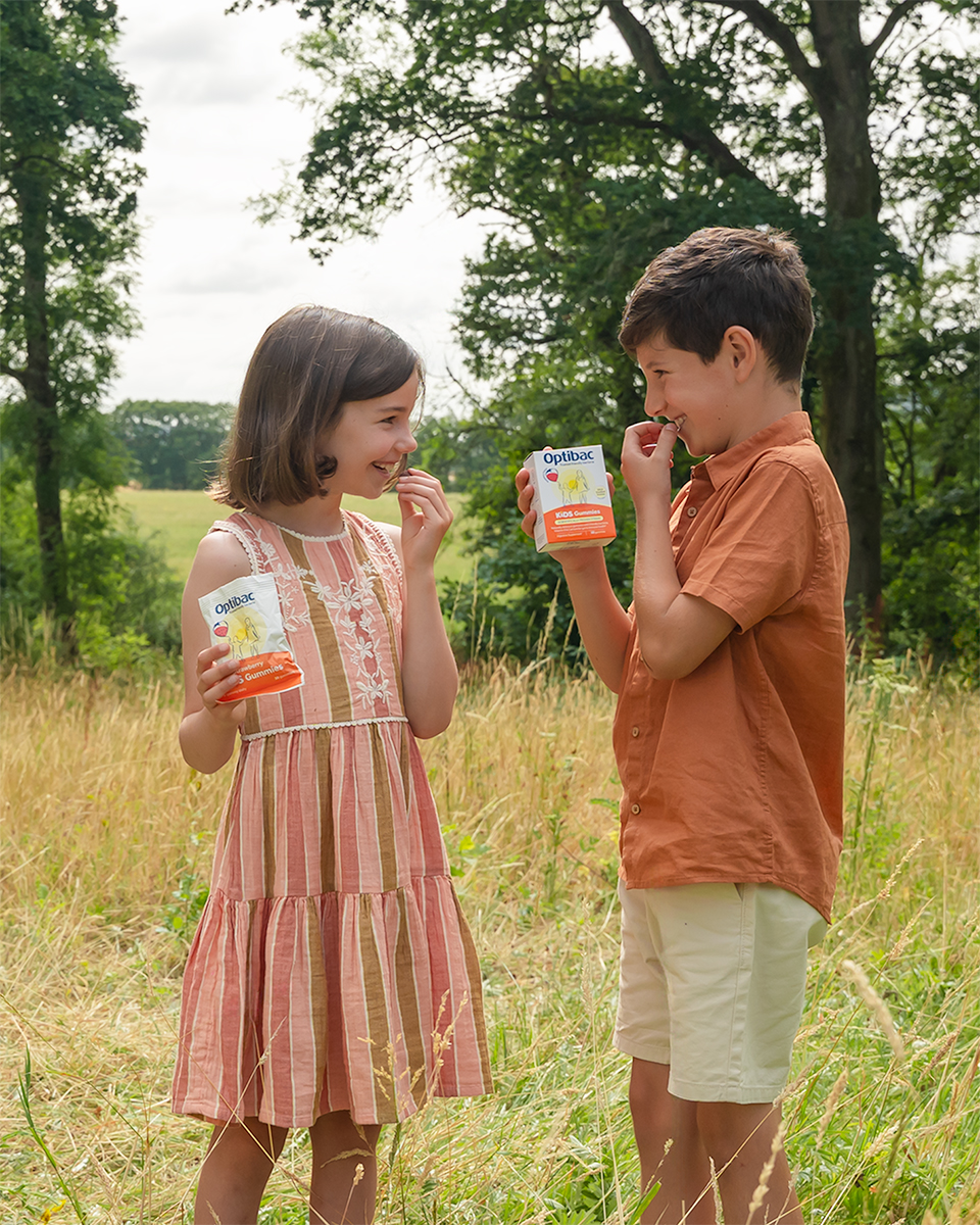 Two children stand in a grassy field, smiling and sharing Optibac Kids Gummies in colorful packaging. Trees and sky are visible in the background.