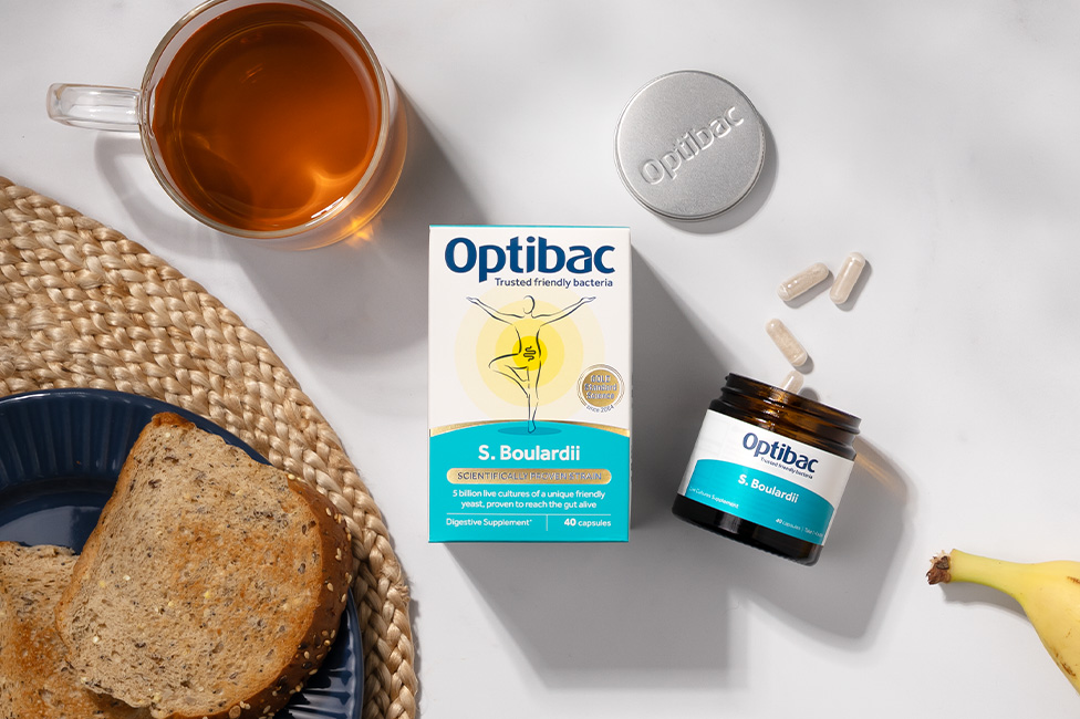 Optibac supplement box and jar on a table with a cup of tea, two capsules, and slices of bread on a blue plate.