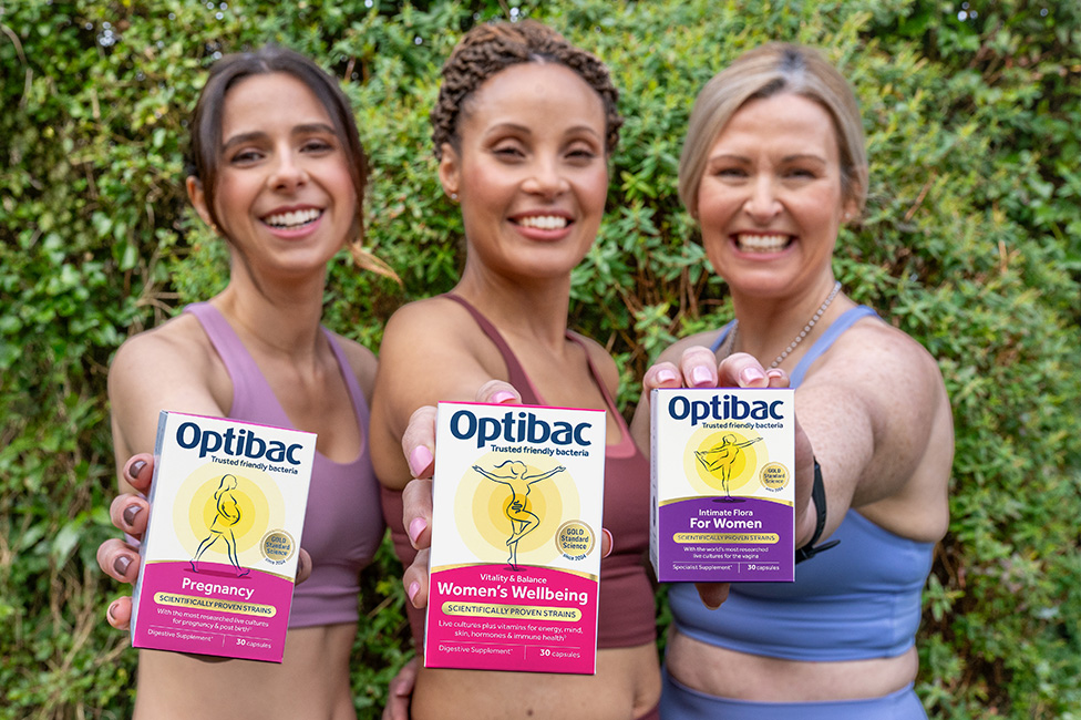 Three women smiling and holding Optibac probiotic boxes in front of greenery, each box labeled for different women's health needs.