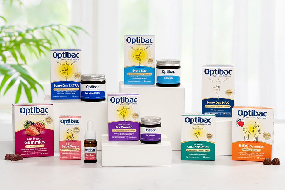 A variety of Optibac probiotic products displayed on white platforms, including gummies, drops, and supplements, with a leafy background.