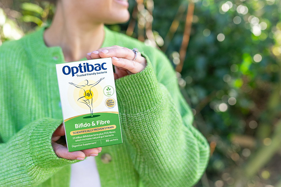 Person in a green sweater holding a box of Optibac Bifido & Fibre probiotic supplement outdoors.