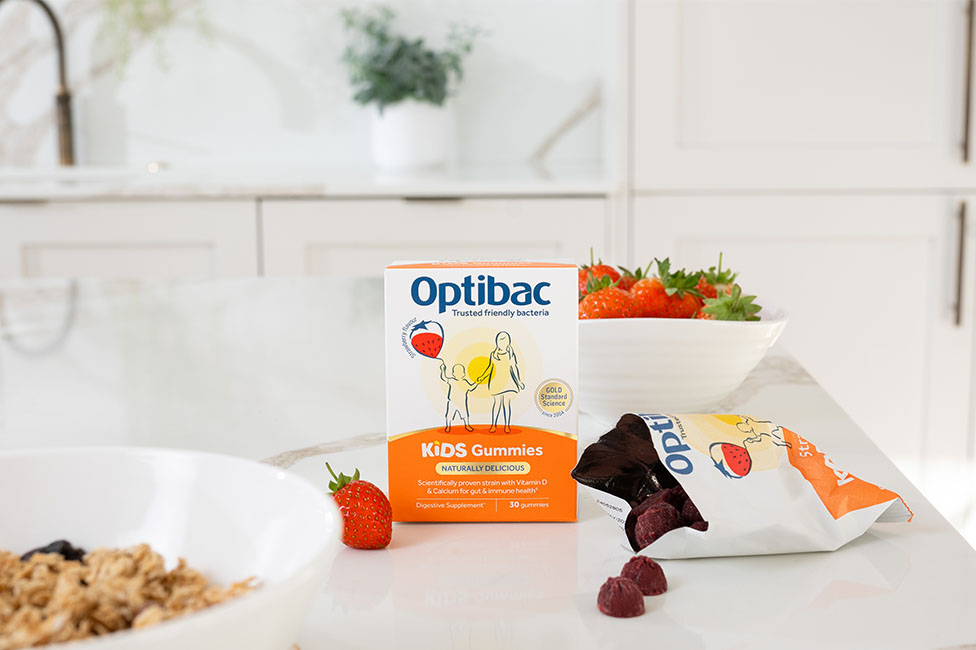 Optibac Kids Gummies box and open pouch on a kitchen counter with strawberries and a bowl of cereal nearby.