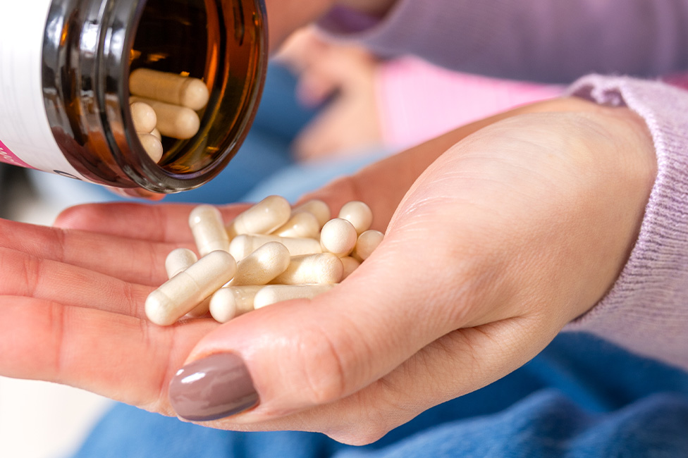 A person pours white capsules from a brown bottle into their hand, wearing a light purple sleeve and nude nail polish.