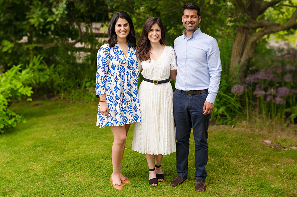 Three people standing on grass, smiling. One wears a blue and white dress, another a white dress, and the third a light blue shirt and dark pants.