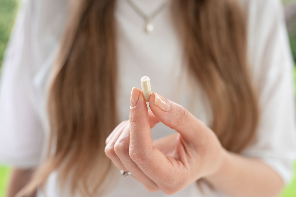 A person with long hair holds a small white capsule between their fingers, wearing a white shirt and a necklace.