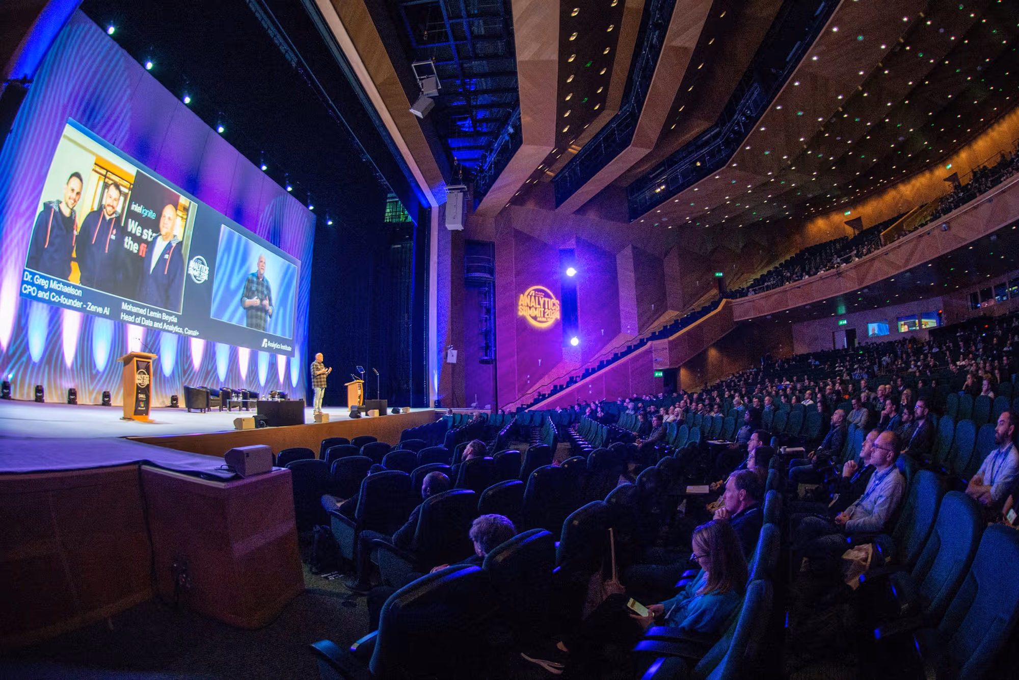 A speaker presents to a full audience at the Data Analytics Summit, highlighting advancements in data and AI applications on a large stage with immersive visuals and lighting.