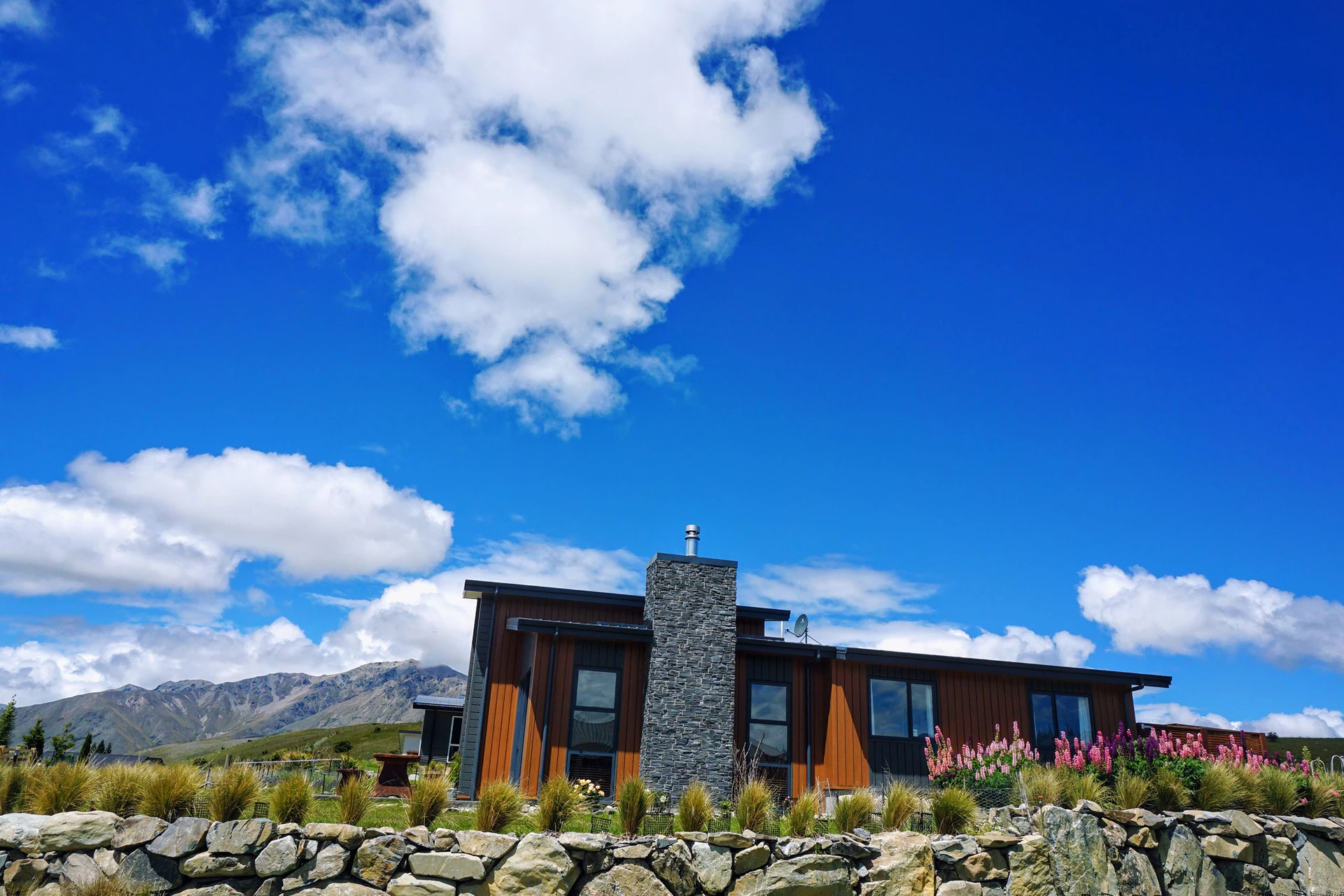 Modern house with stone chimney, surrounded by a stone wall and flowers, set against a bright blue sky and distant mountains.